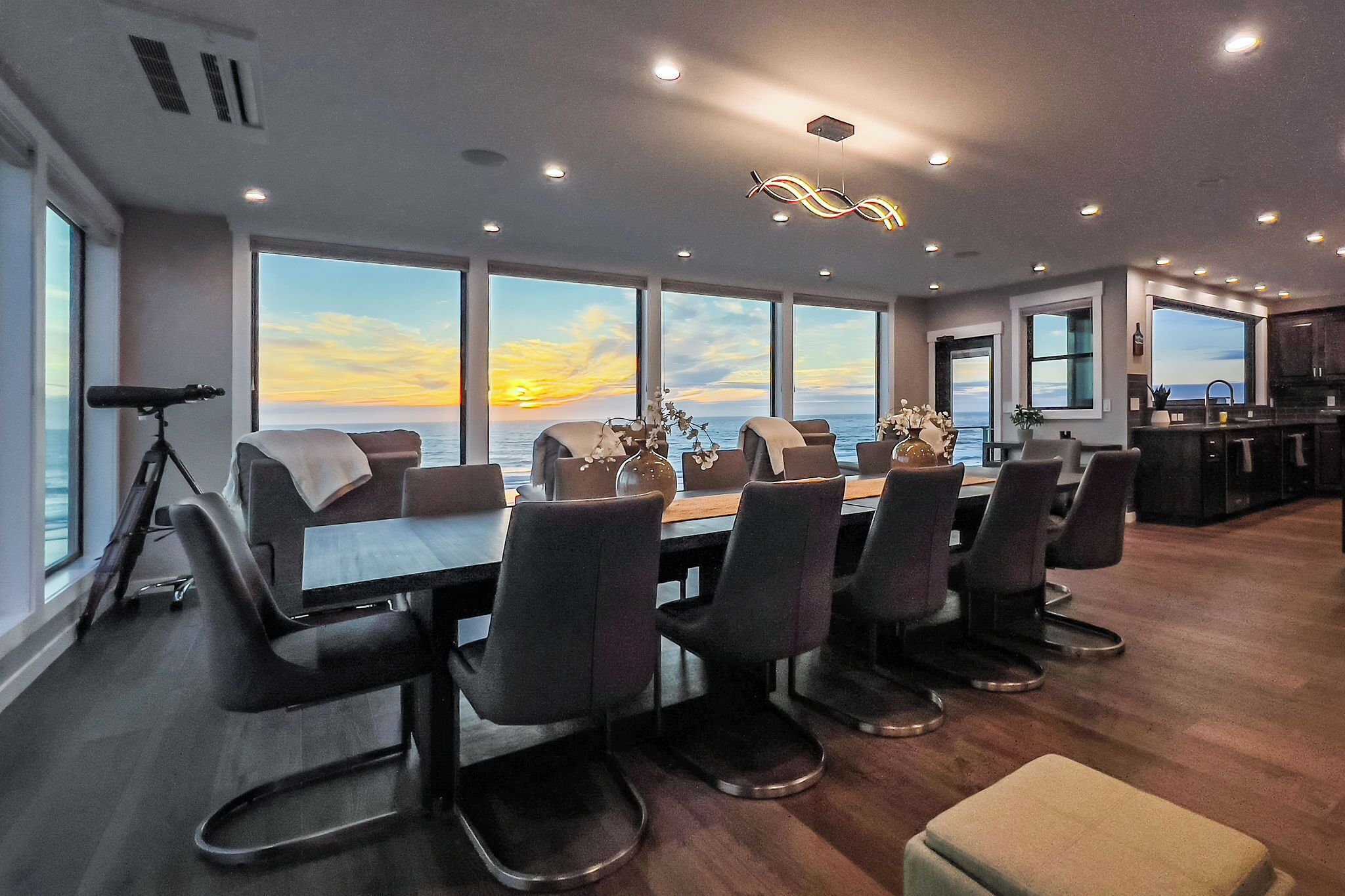 A modern dining area with a view of the sunset over the ocean, a telescope, and a kitchen in the background.