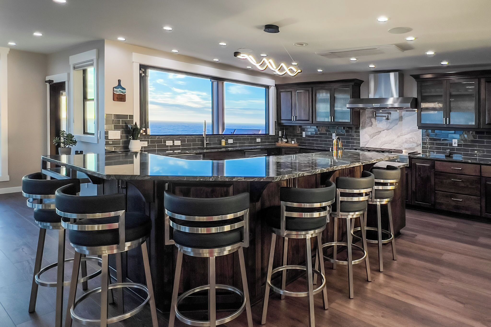 A modern kitchen with a large island, bar stools, dark cabinets, and a view of the ocean.