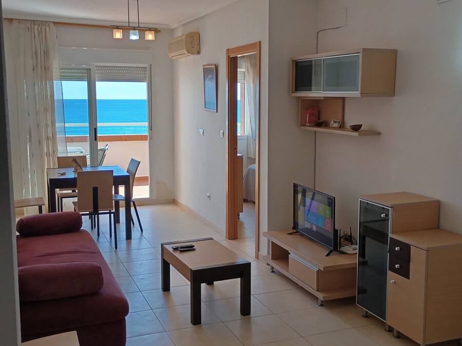 A living room with a sofa, a coffee table, a TV, and a kitchenette. There is a view of the ocean through a glass door.