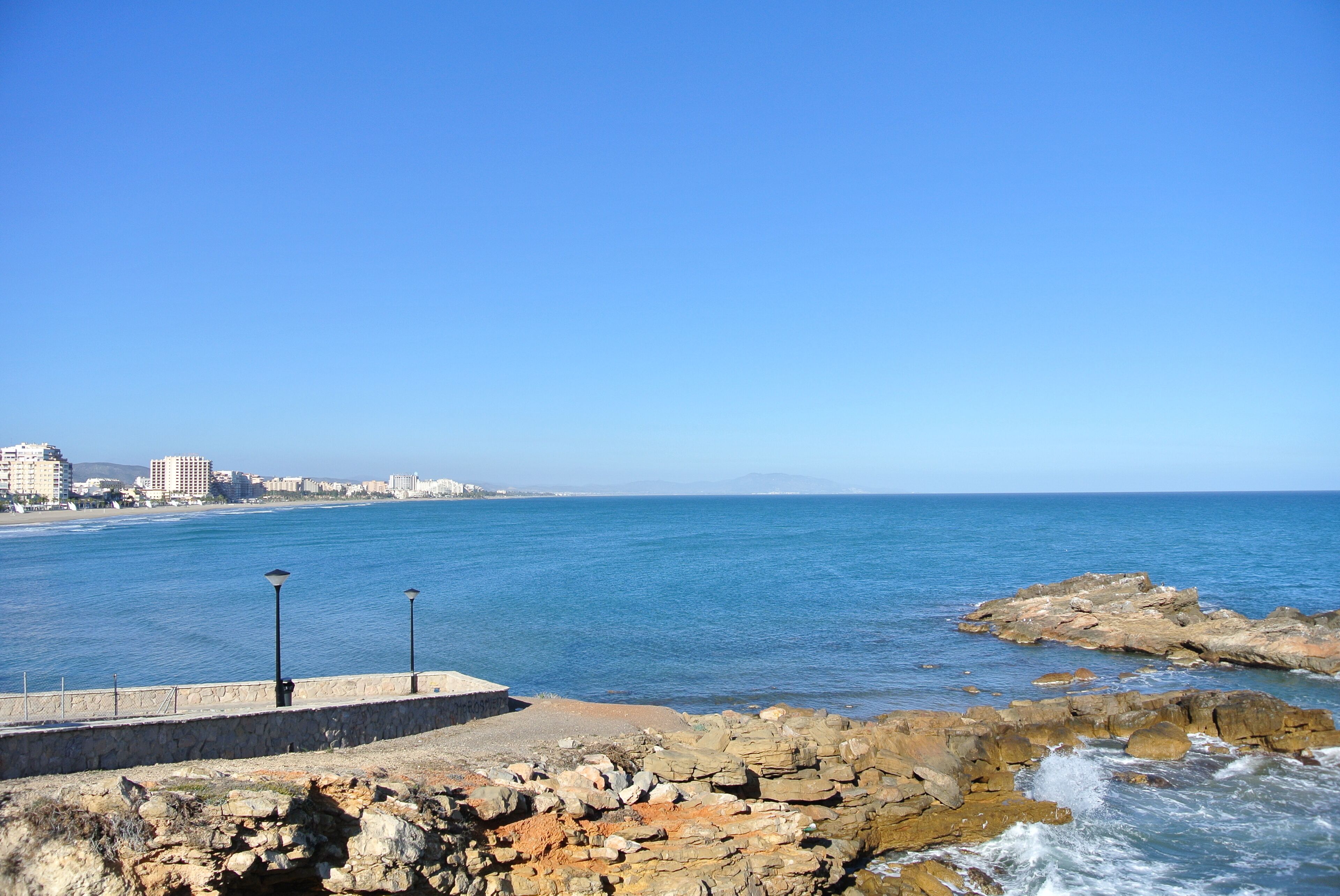 Vista costera con muelle de piedra, costa rocosa y edificios a lo lejos.