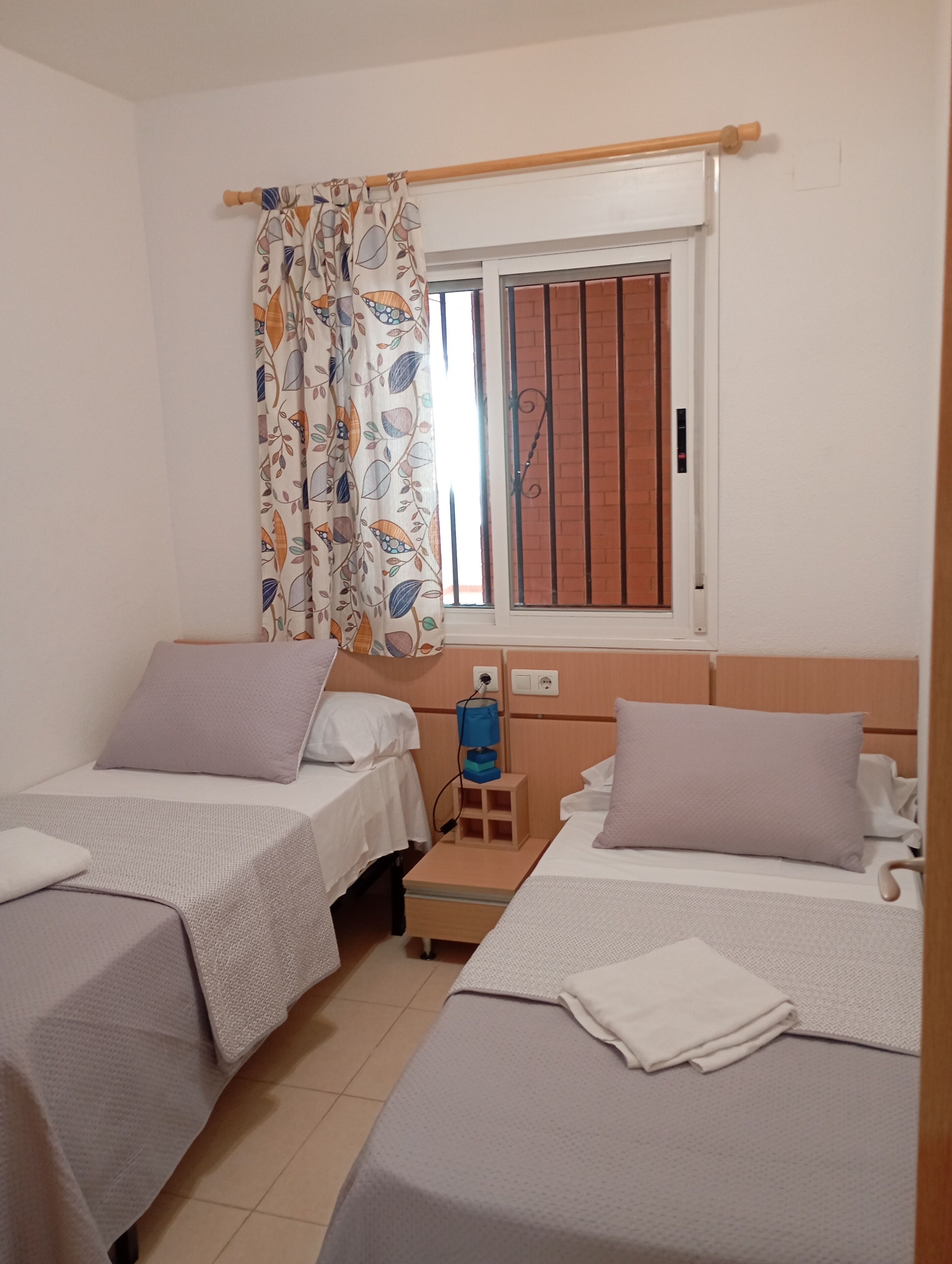 Habitación doble con dos camas, una ventana con cortina, una mesita de noche con lámpara y un enchufe en la pared.