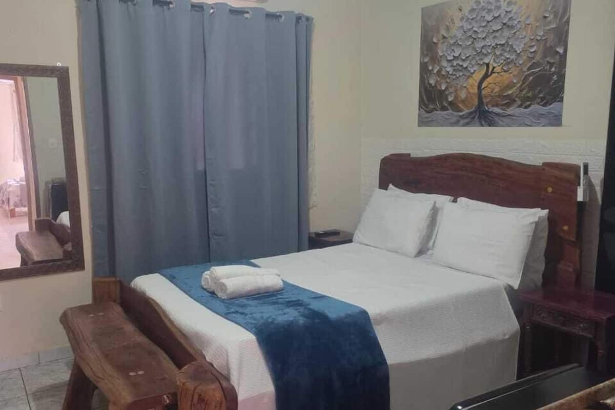 A bedroom with a bed, a bench, an air conditioner, a painting, and a refrigerator.