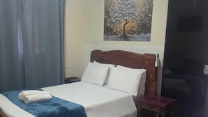 A hotel room with a bed, a nightstand, a desk with a computer, a chair, a suitcase, and a painting on the wall. - Suites Boa Viagem (Pirenópolis)