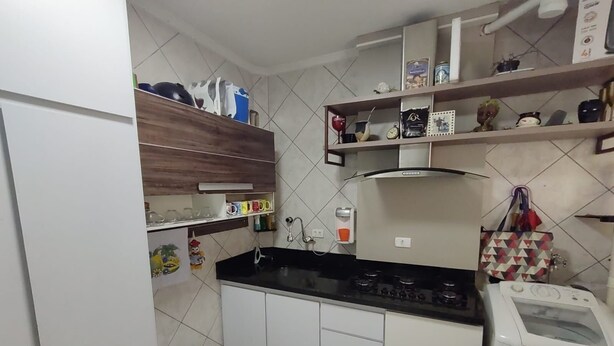 A compact kitchen with white cabinets, a black countertop, and a built-in oven. There are shelves above the sink holding various kitchen items and a trash bin to the left.