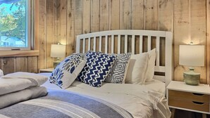 Une chambre avec un mur lambrissé en bois, un lit blanc agrémenté d’oreillers à motifs, une table de chevet avec une lampe et une fenêtre donnant sur des arbres.