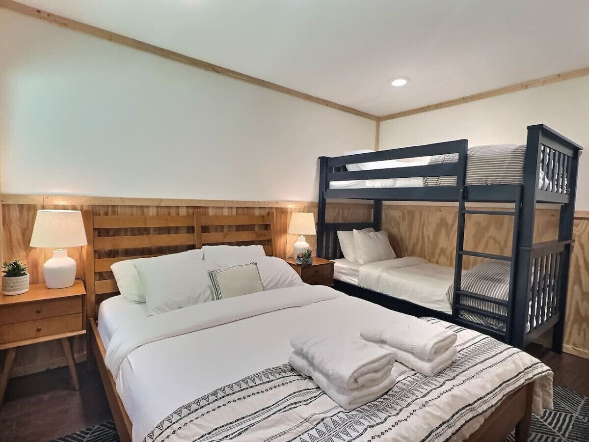 A bedroom with a bunk bed, a single bed, wooden headboard, bedside tables with lamps, and a patterned bedspread.