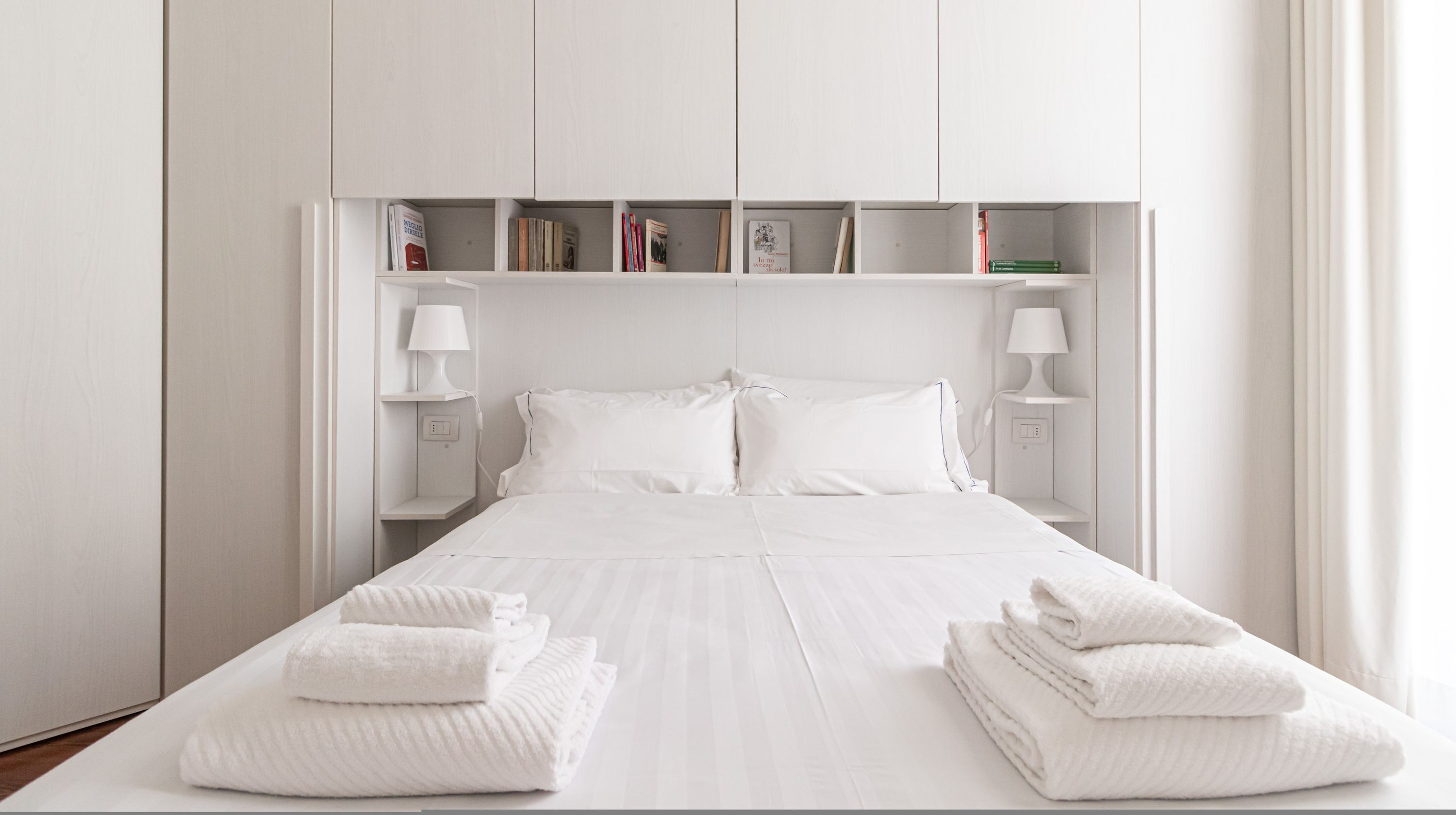 A neatly made bed with white linens and towels, a bedside lamp, and a shelf with books and a small lamp.