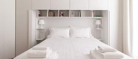Un lit bien fait, avec du linge de lit et des serviettes blanches, une lampe de chevet et une étagère avec des livres et une petite lampe.