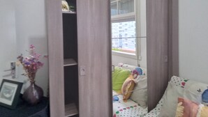 A room with a sliding wardrobe, a window with a view of buildings, a framed picture on the left, and a vase with flowers.