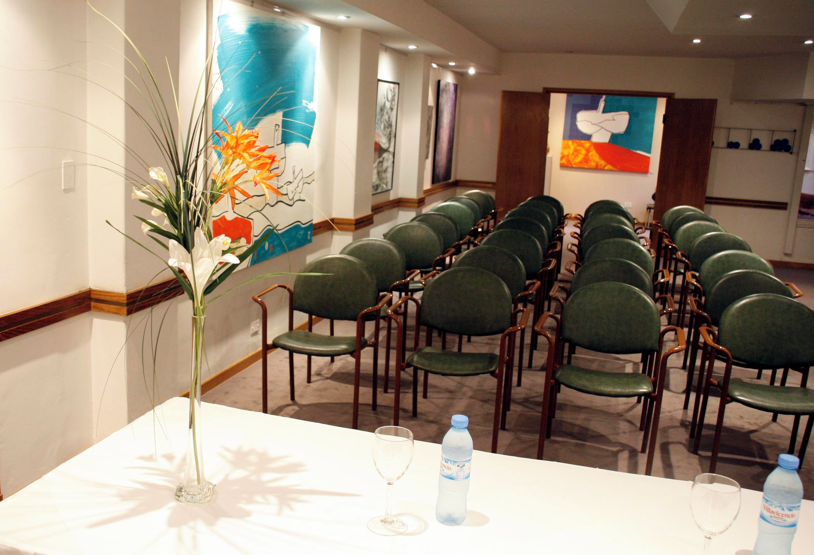 Een vergaderzaal met rijen groene stoelen, een witte tafel en een vaas met bloemen.