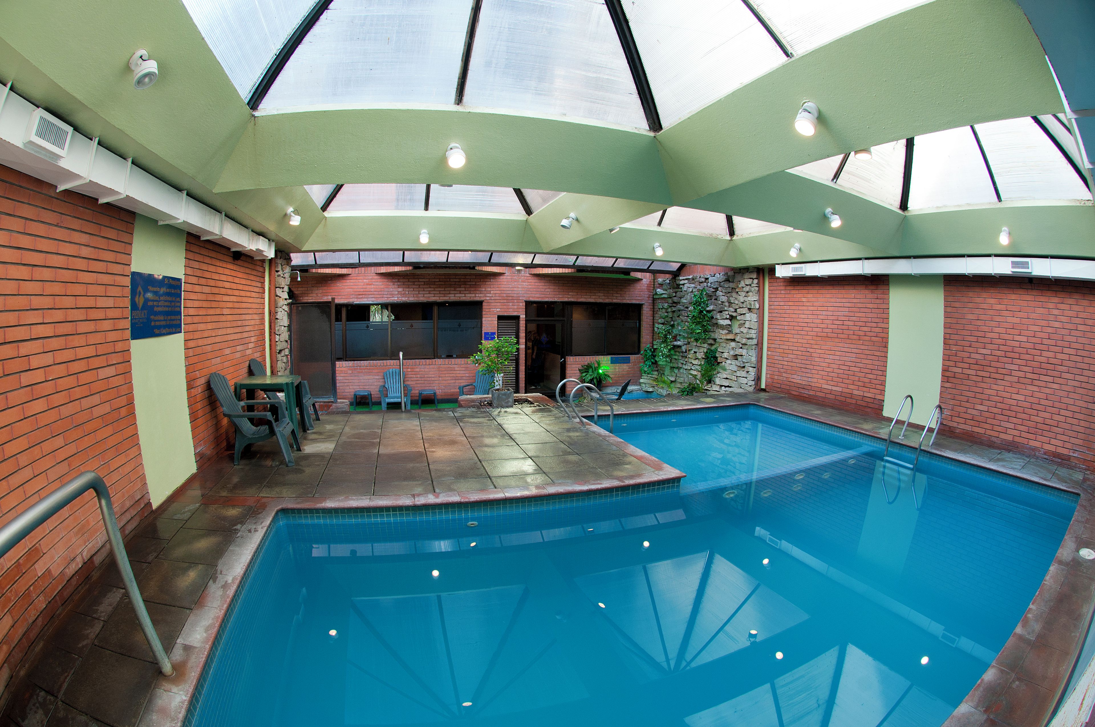 An indoor swimming pool with a glass ceiling, brick walls, and a seating area with chairs.