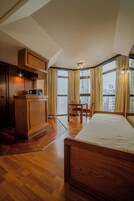 Habitación de hotel con cama de madera, kitchenette, zona de comedor y ventanas grandes con cortinas.