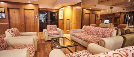 Un vestíbulo de hotel con sofás y sillones, una mesa de centro de vidrio, puertas de madera y un televisor.