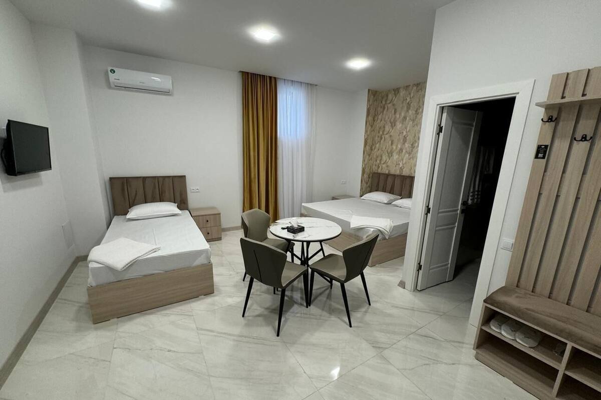 Habitación de hotel con dos camas individuales, una mesa redonda con sillas, un televisor montado en la pared y un armario con estante para zapatos.