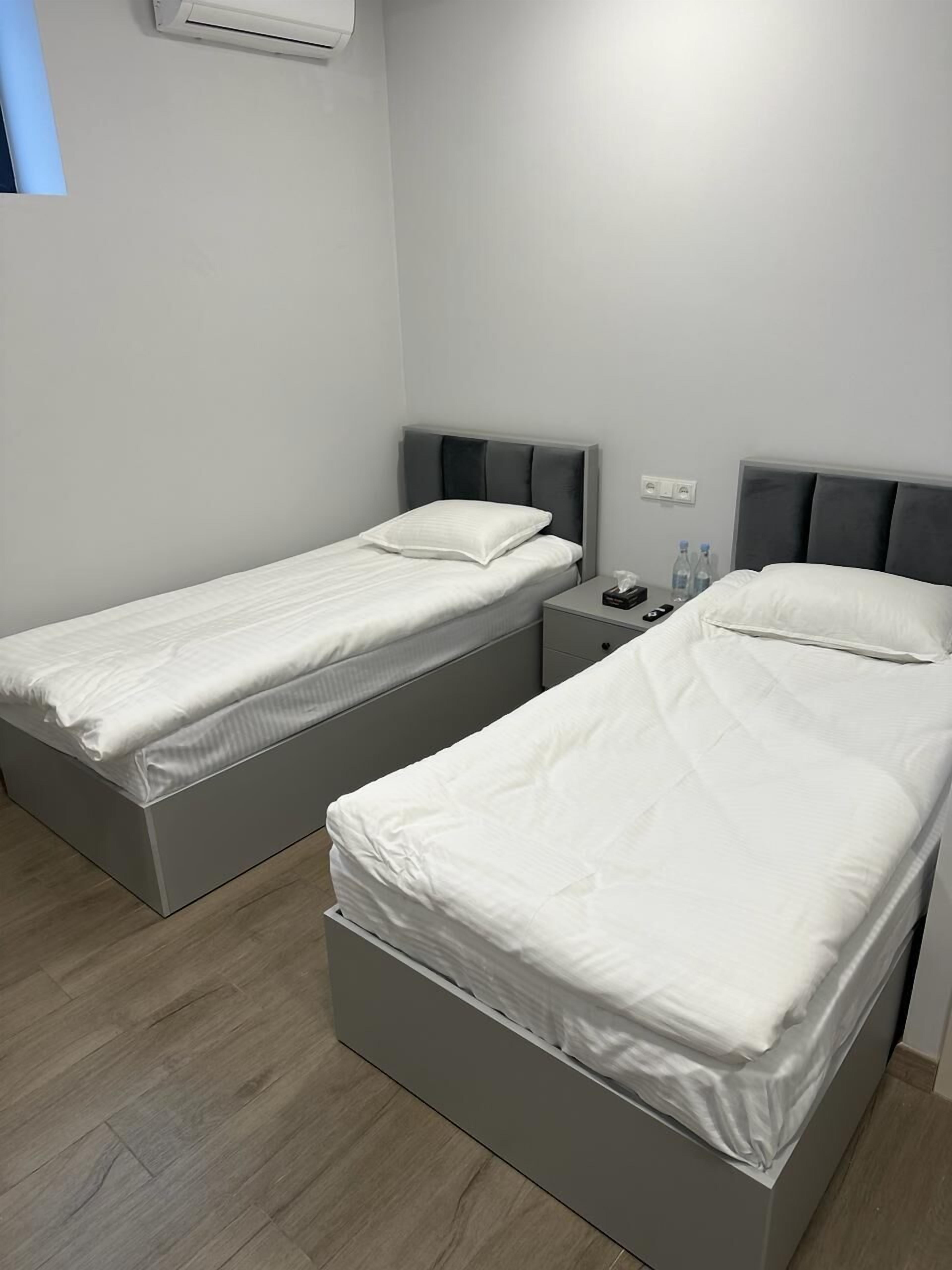 A room with two single beds, a bedside table with a lamp, and a wall-mounted air conditioner.