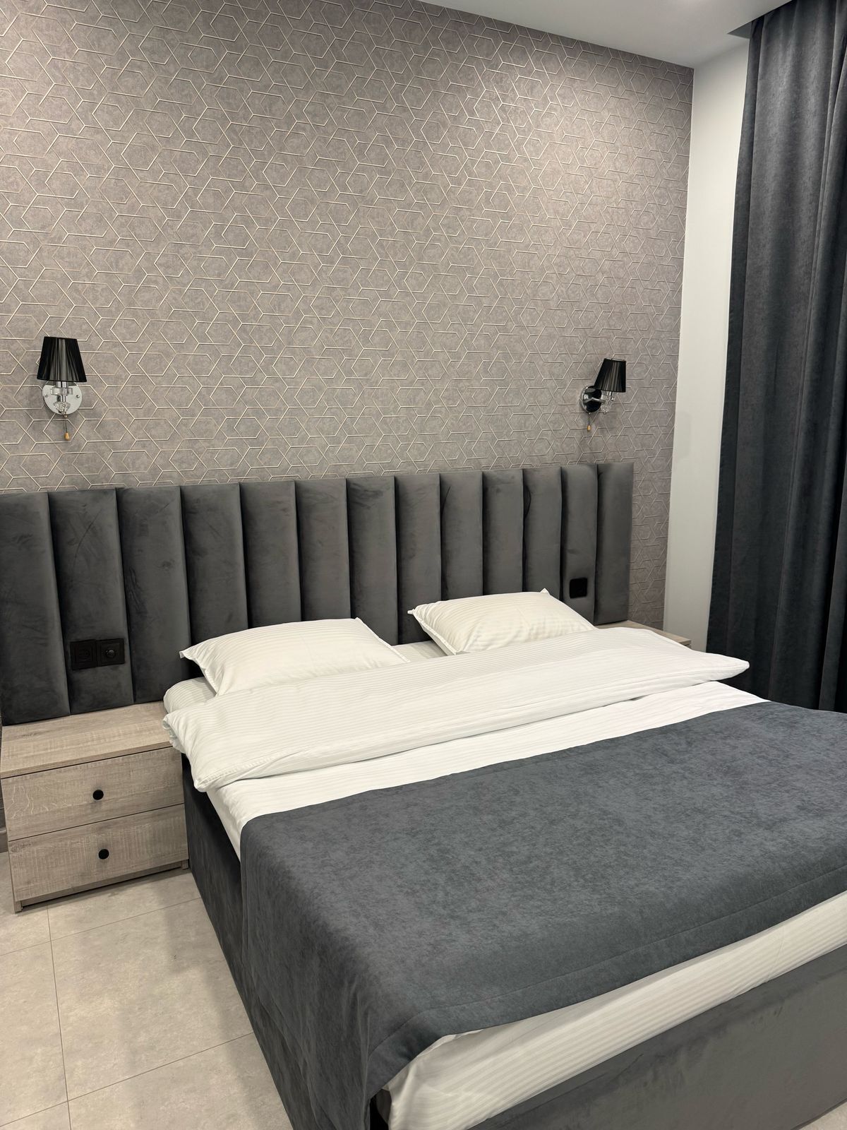 Een moderne slaapkamer met een wand met een geometrisch patroon, een grijs gestoffeerd hoofdbord, twee nachtkastjes met lampen en een grijze dekbedovertrek.