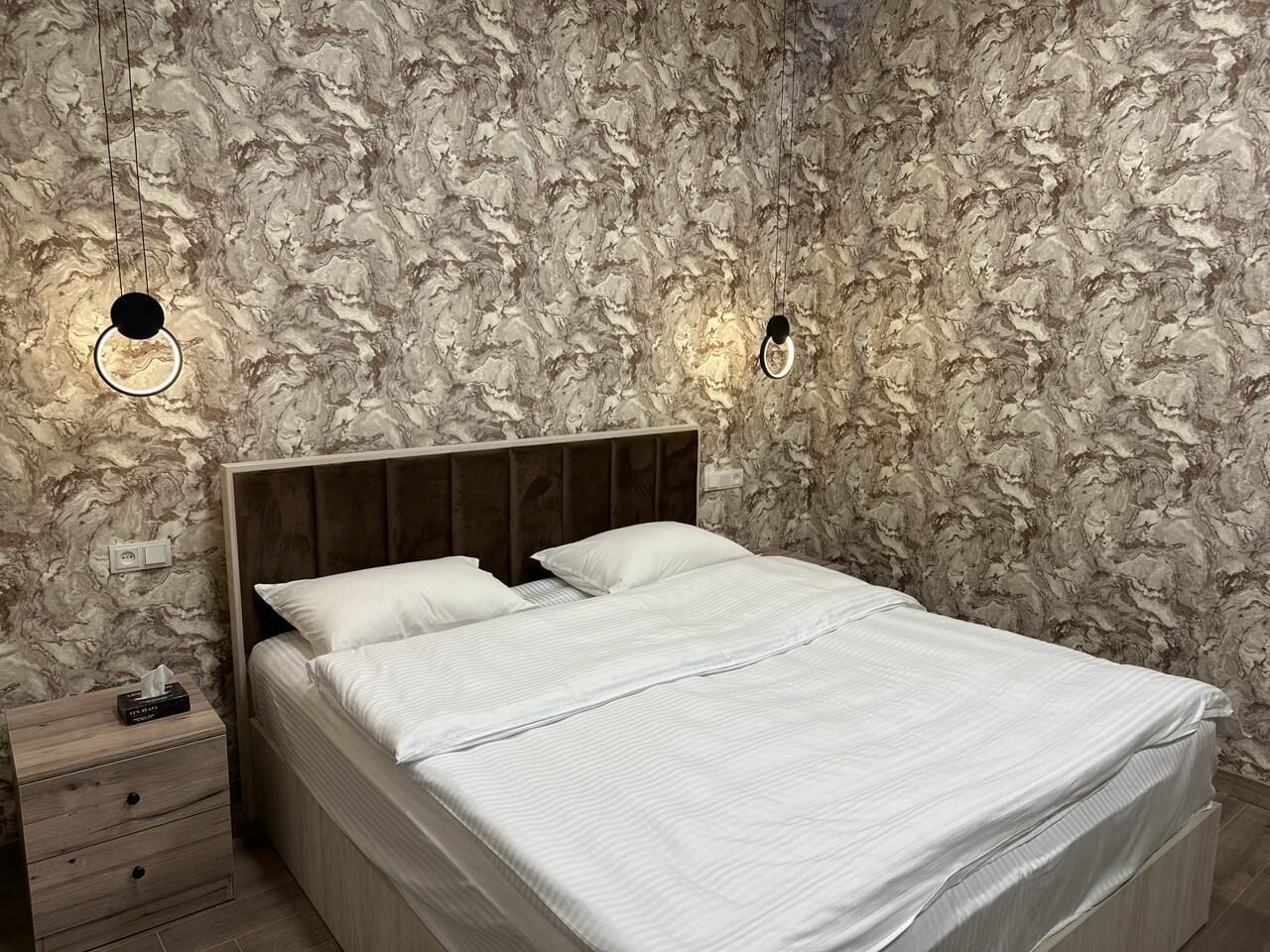 A bedroom with a marble-patterned wall, a wooden headboard, a bed with white bedding, and two wall-mounted light fixtures.