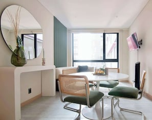 A modern dining area with a round table, green upholstered chairs, a large mirror, and a TV mounted on the wall.