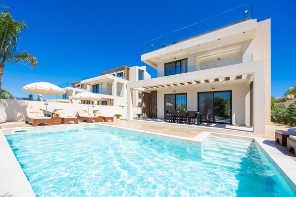 A modern house with a swimming pool, outdoor seating, and a clear blue sky.