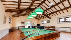 Une table de billard avec des billes, une queue de billard et un luminaire vert suspendu dans une pièce aux poutres apparentes et au sol carrelé.