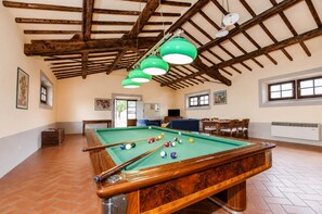 Una mesa de billar con bolas, una vara de billar y una lámpara colgante verde en una habitación con vigas de madera y suelo de baldosas.
