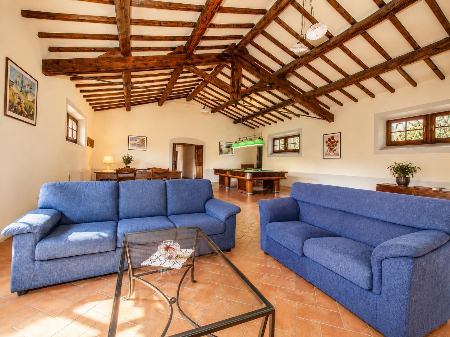 A spacious living room with two blue sofas, a glass coffee table, a pool table, and wooden beams on the ceiling.