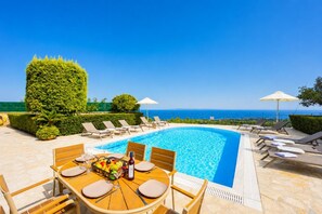 Un área de comedor junto a la piscina, con una mesa dispuesta para dos personas, rodeada de sillones de descanso y sombrillas, con vistas a un cielo azul claro y al océano.