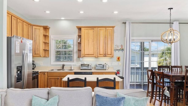 A modern kitchen with wooden cabinets, a stainless steel refrigerator, a dining area with a table and chairs, and a large window providing natural light.
