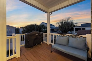 A covered outdoor deck with a grill, a cushioned bench, and a view of neighboring houses.