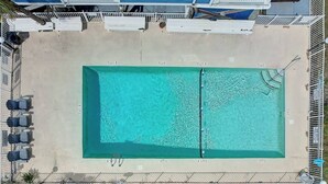 Vista aérea de una piscina rectangular con zona de terraza, rodeada por una valla y protegida por sombrillas.