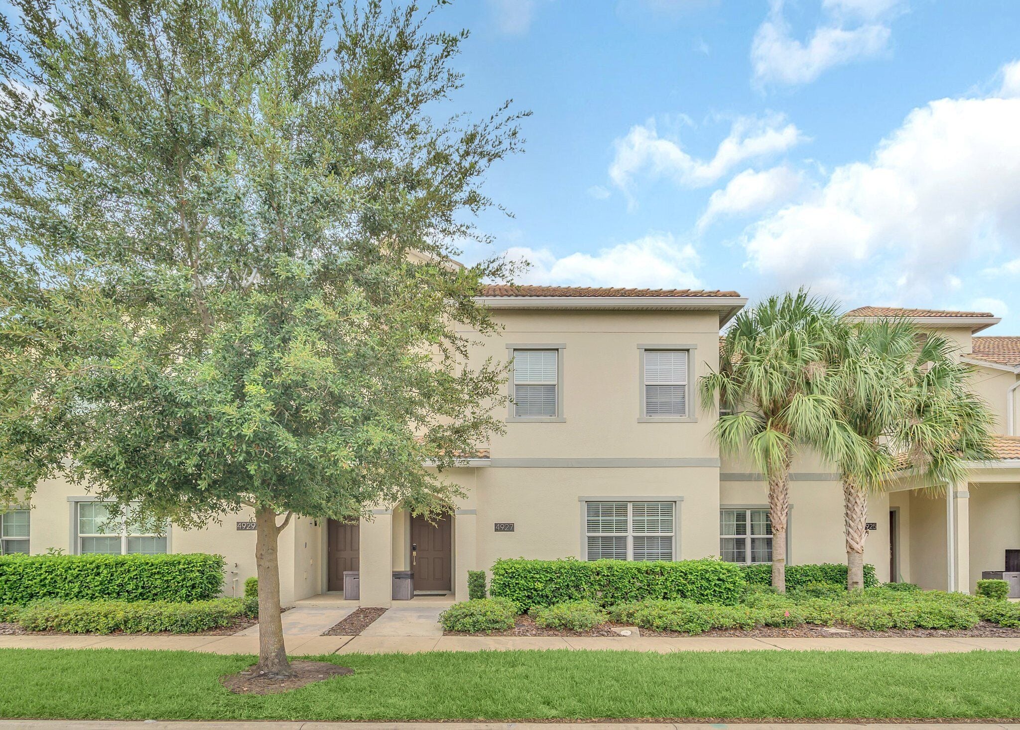 A two-story residential building with a beige facade, multiple windows, and a well-maintained lawn with a large tree and palm trees.