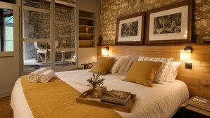 Camera d'hotel con un letto grande, una testiera in legno, due lampade a parete, un vassoio con un libro e un vaso, e una parete in pietra con quadri appesi.