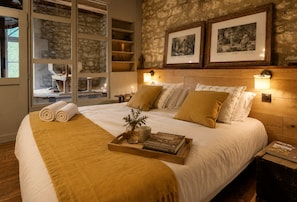 A hotel room with a large bed, a wooden headboard, two wall-mounted lamps, a tray with a book and a vase, and a stone wall with framed pictures.