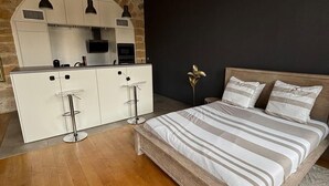A modern bedroom with a wooden bed, a white dresser, and a kitchen area with a stone backsplash. - The panorama (Lyon)