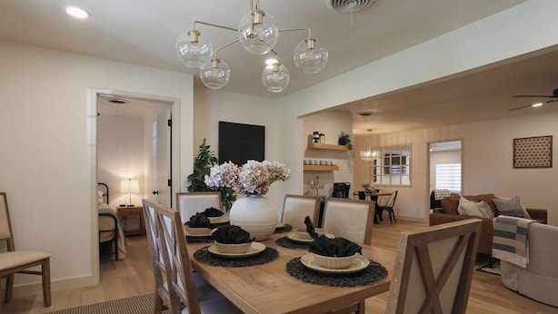 Un coin repas avec une table en bois, dressée pour quatre personnes, entourée de chaises. Un vase rempli de fleurs est posé sur la table, et un luminaire est suspendu au plafond.