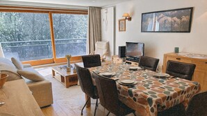 A dining area with a table set for four, a television, and a view of trees through large windows.