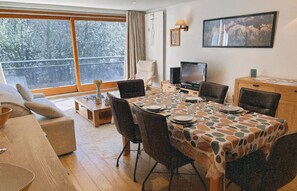 Un coin repas avec une table dressée pour quatre personnes, un téléviseur et une vue sur les arbres à travers de grandes fenêtres.