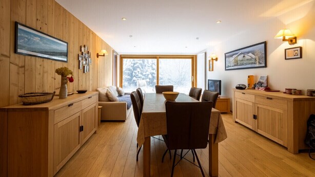 A modern kitchen with wooden cabinetry, a dining table with chairs, and a large window with a view of snow-covered trees.