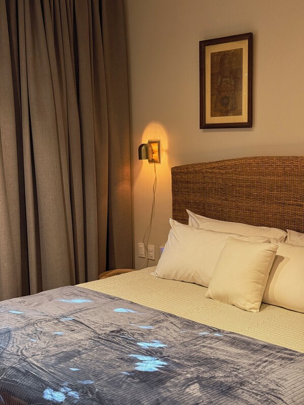 Ett hotellrum med en säng, en vägglampa, en inramad tavla och ett fönster med gardiner.