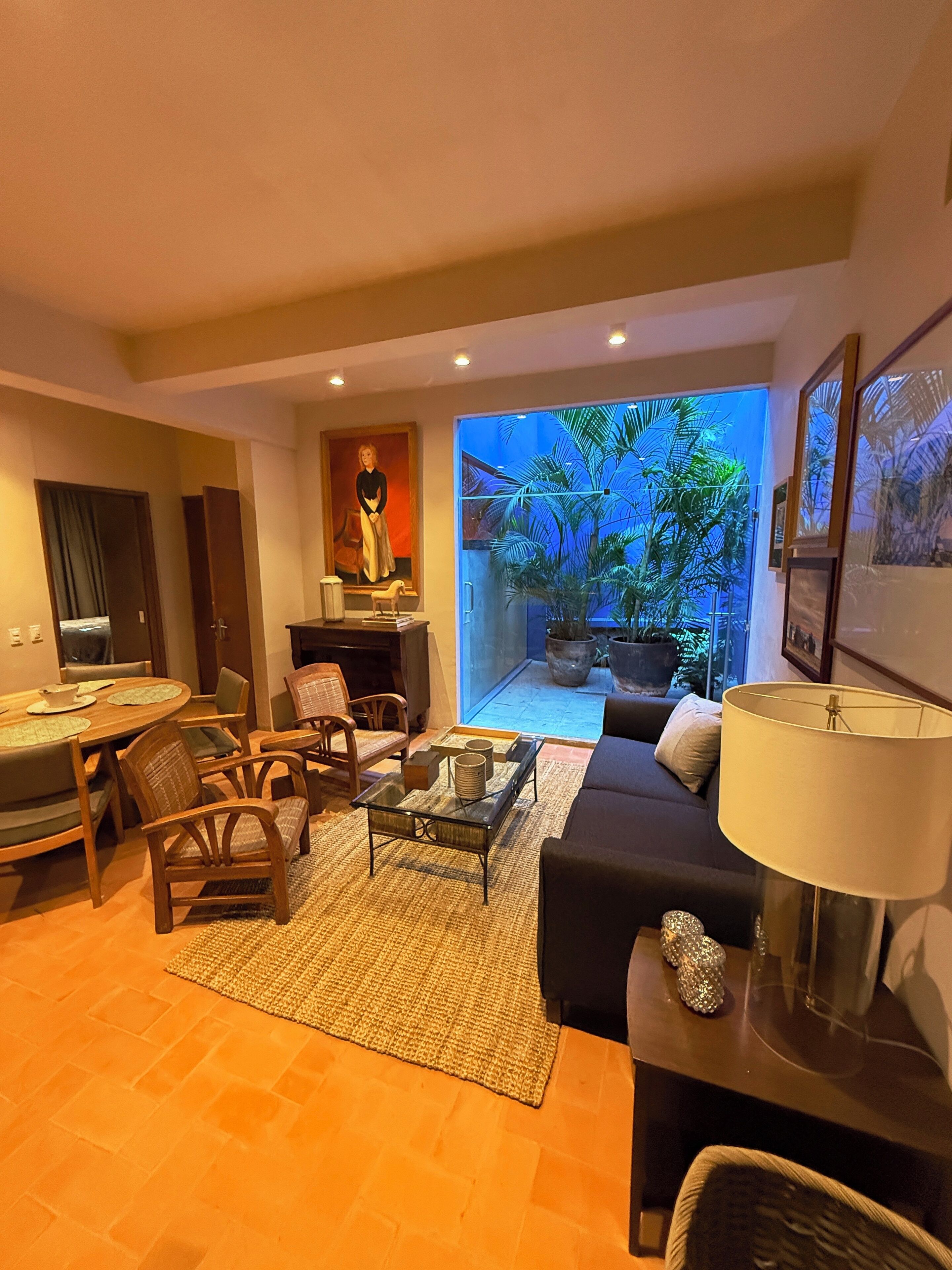 Una sala moderna con comedor, sofá, mesa de centro y vistas a un jardín tropical a través de un ventanal.