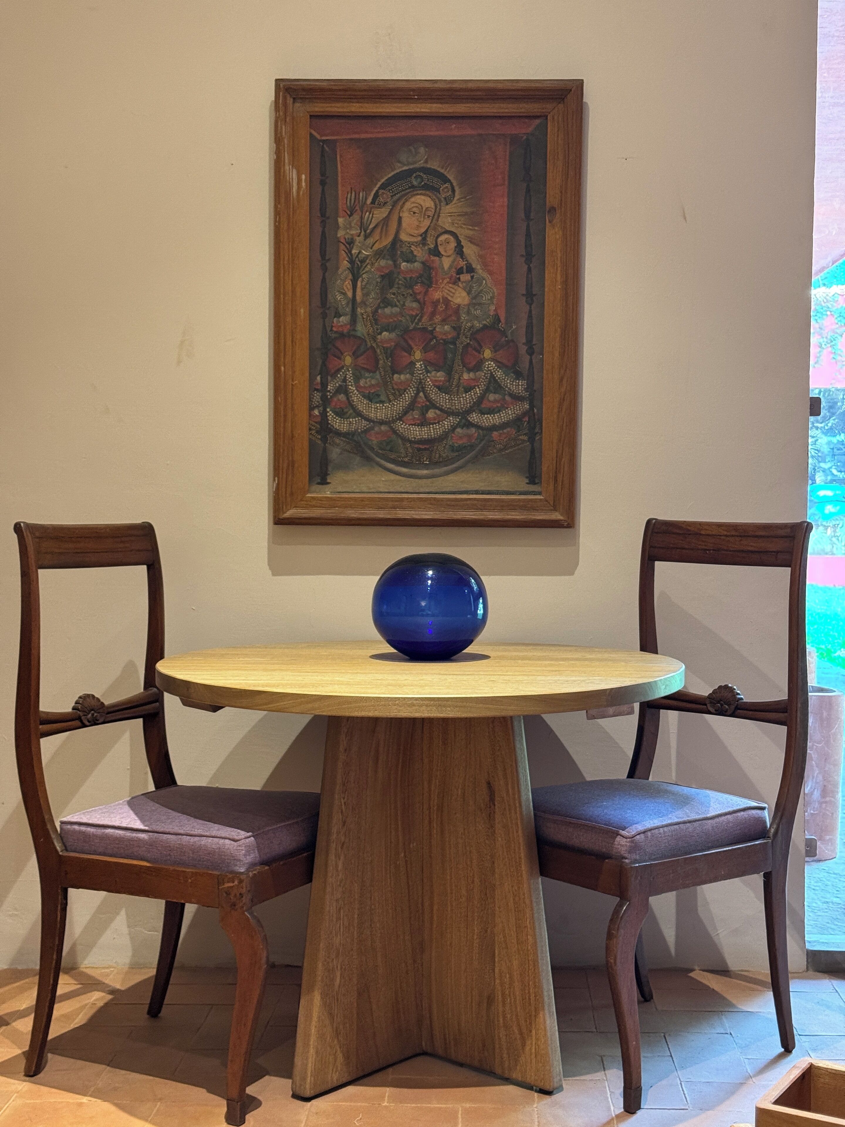 A dining table with a blue vase, two wooden chairs, and a framed painting above.