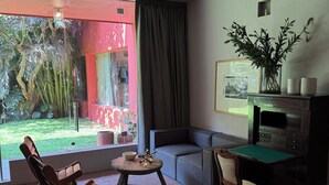 A modern living room with a large window, a sofa, a wooden chair, a small table, and a desk with a plant. - Parque Juan Diego (Zapopan)
