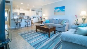 A modern living room with a grey sofa, a coffee table, and a striped rug. There is a kitchen area with white cabinets and a dining area with bar stools. - Sunsets over Siesta Key in this cozy 2+2 condo
 (Sarasota)