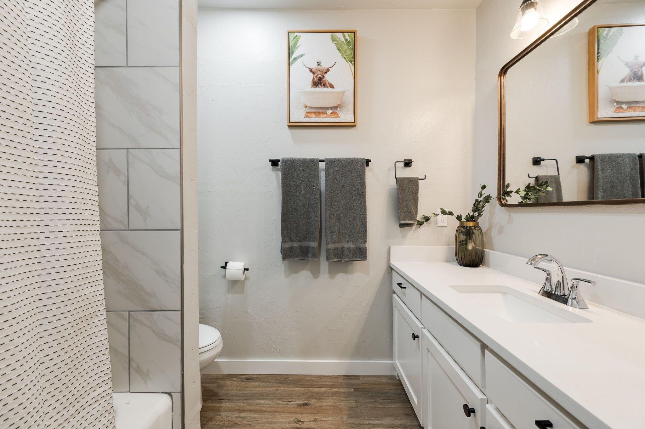 A modern bathroom with a white sink, a mirror, and a toilet. There are two towels hanging on hooks, and a framed picture of a dog in a bathtub on the wall.