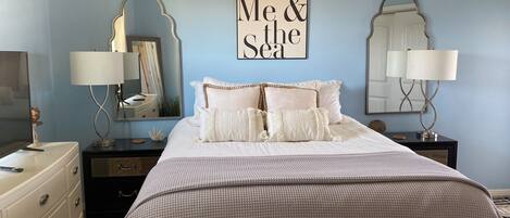 A bedroom with a bed, two nightstands, a dresser, and a mirror. There is a wall-mounted sign that reads "You Me & the Sea".