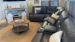 Una sala de estar moderna con un sofá de cuero, una mesa de centro, un televisor de pantalla plana, decoración en la pared y un amplio ventanal con persianas.