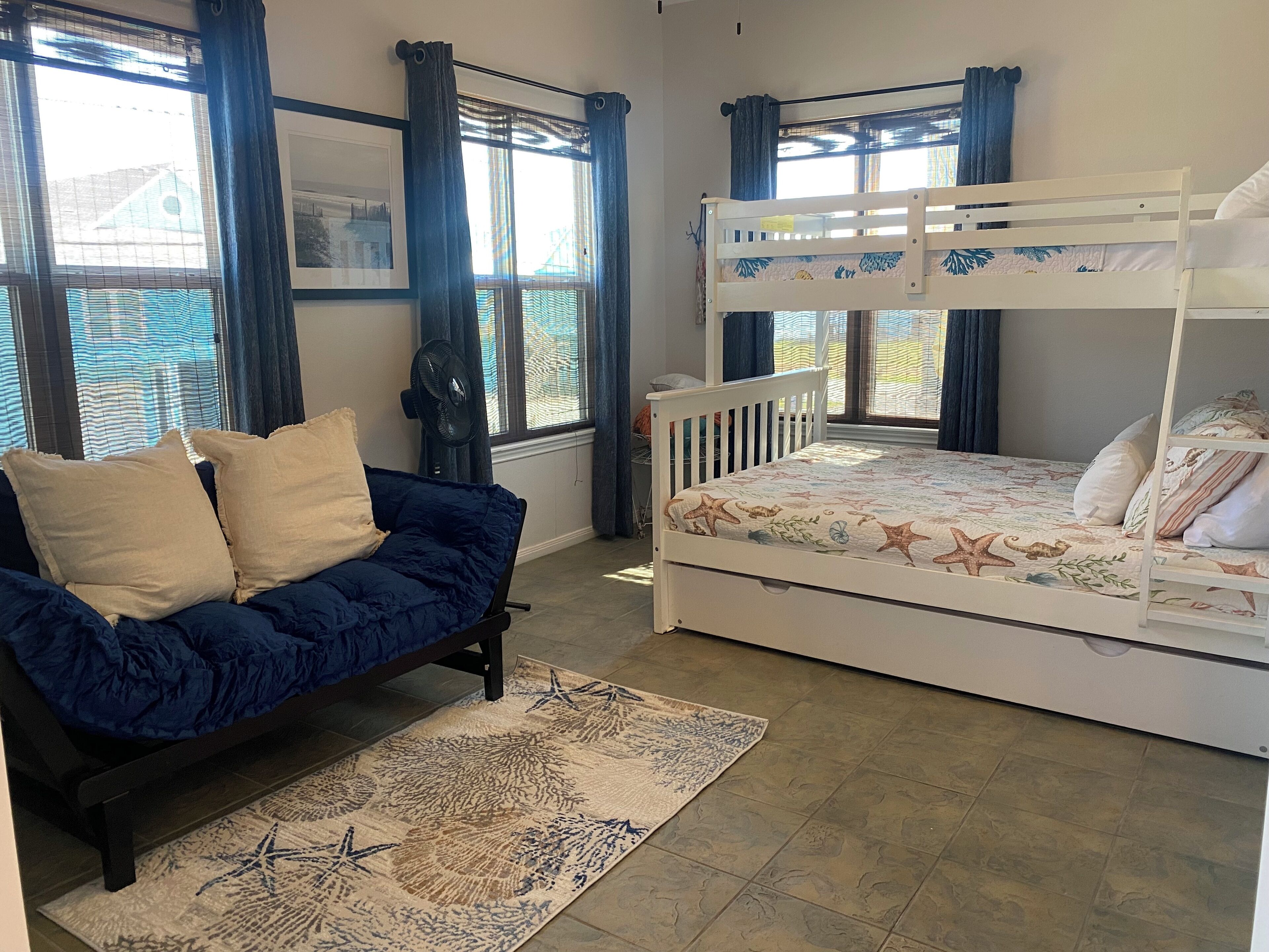 A bedroom with a bunk bed, a sofa bed, a large window with curtains, a fan, and a rug.