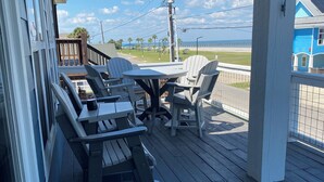 Terraza de madera con mobiliario exterior, mesas y sillas, con vista a una playa y al océano.