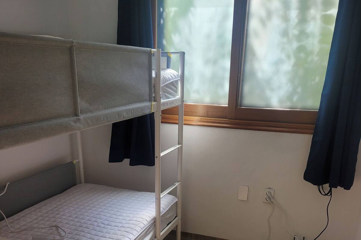 A small, well-lit room with a bunk bed, a window with curtains, and a wooden floor.