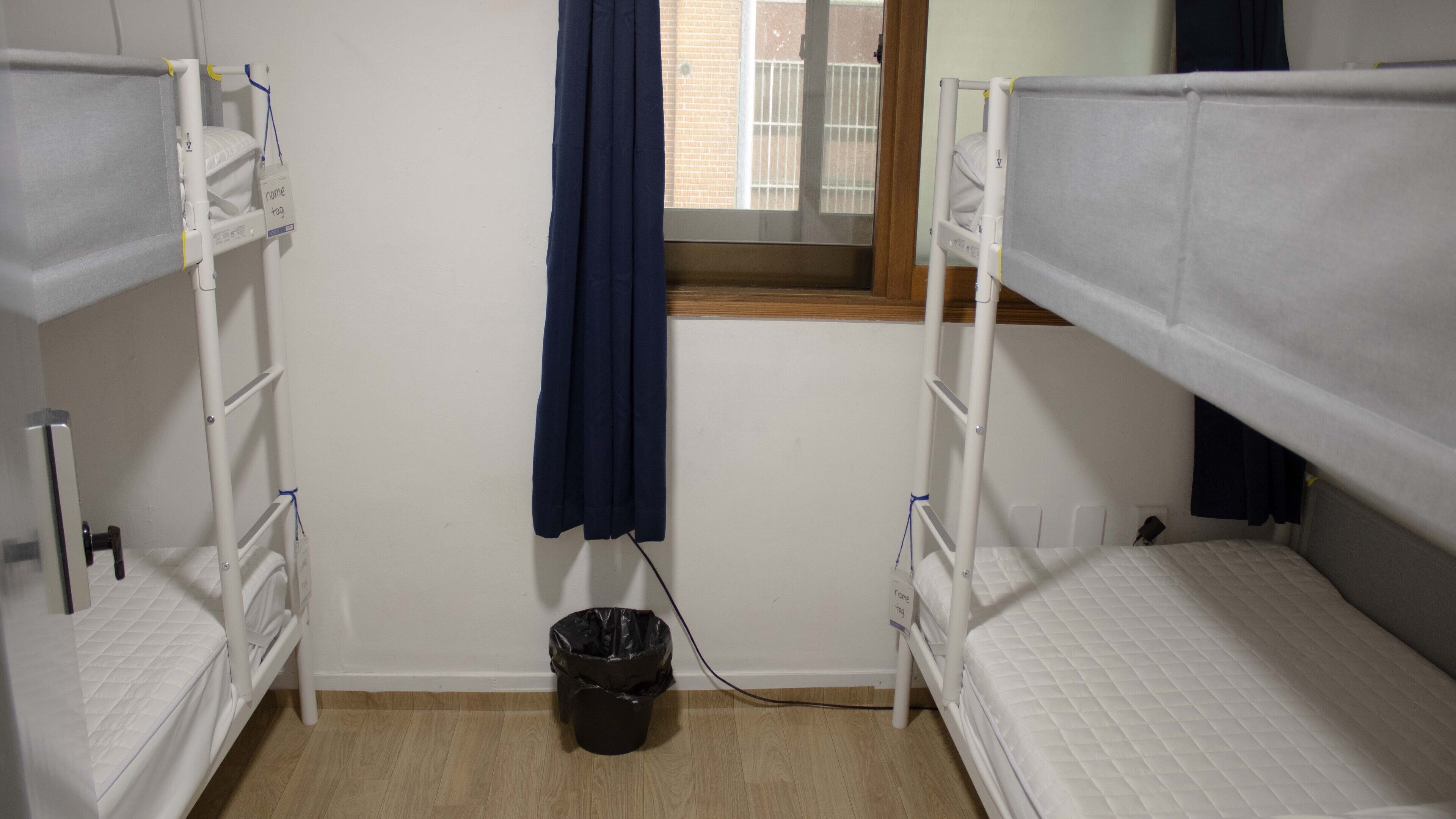 A dormitory room with bunk beds, a window with a view of a brick building, and a black trash bin.